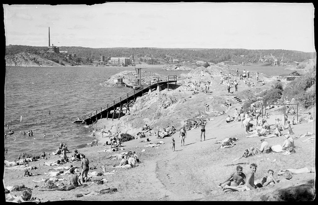 Badplats med solbadande människor på standen. Rutschkanan som syns i mitten av bilden byggdes 1950. TIll höger i bild sitter några pojkar och tittar in i kameran.