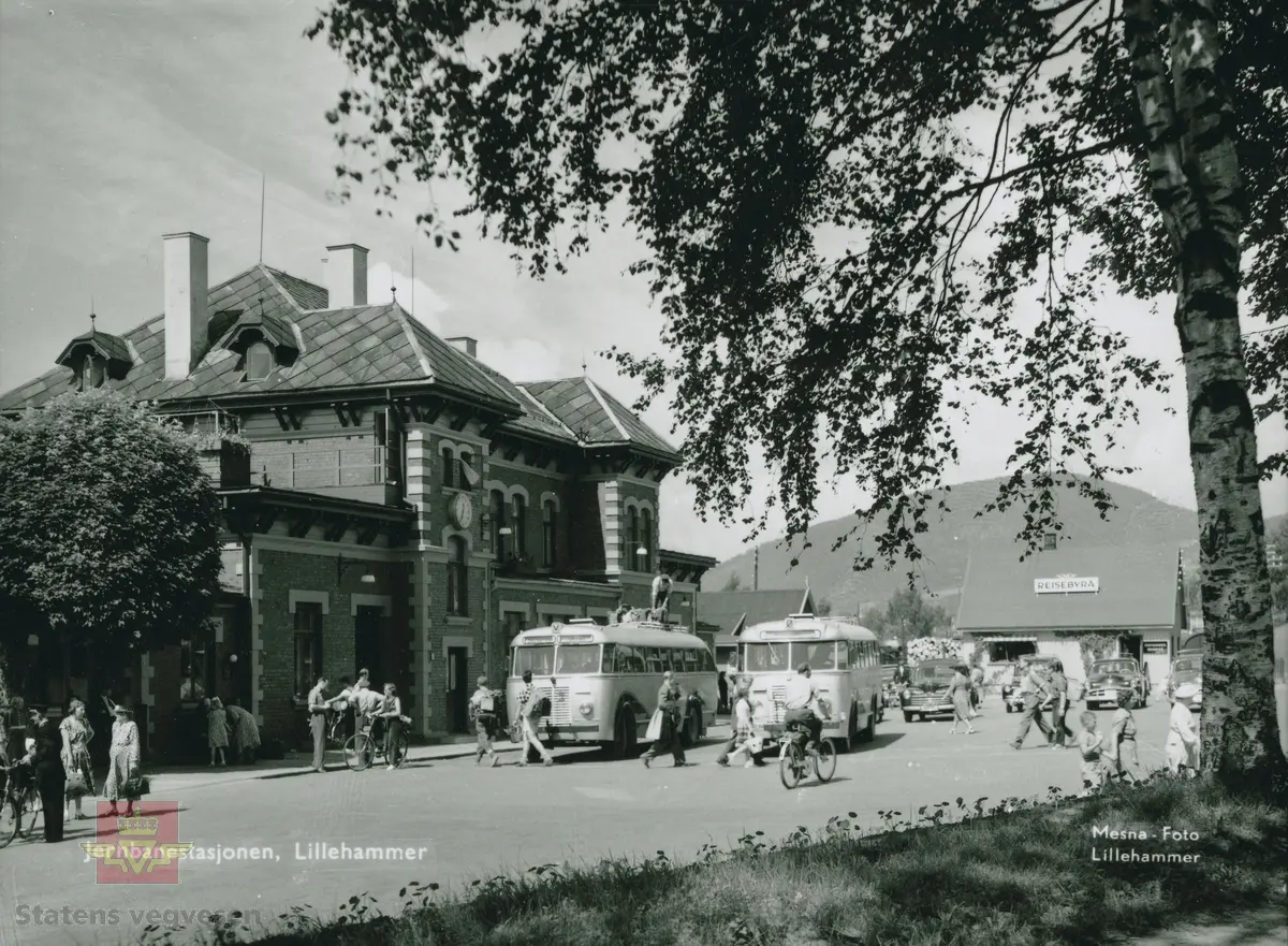 Bilde fra Lillehammer jernbanestasjon en sommerdag 1951-1956, som viser både gående, syklende og reisende til og fra stasjonen. Reisebyrået er til høyre på bildet. Skiltet på bussen til venstre viser Dalseter og Skåbu, hvor buss-sjåføren står på taket og fester bagasjen til passasjerer. Bussen til høyre er skiltet til Gausdal. Bilen ved siden av bussen er en Ford og bilen nr. 3 fra venstre er en Plymouth.