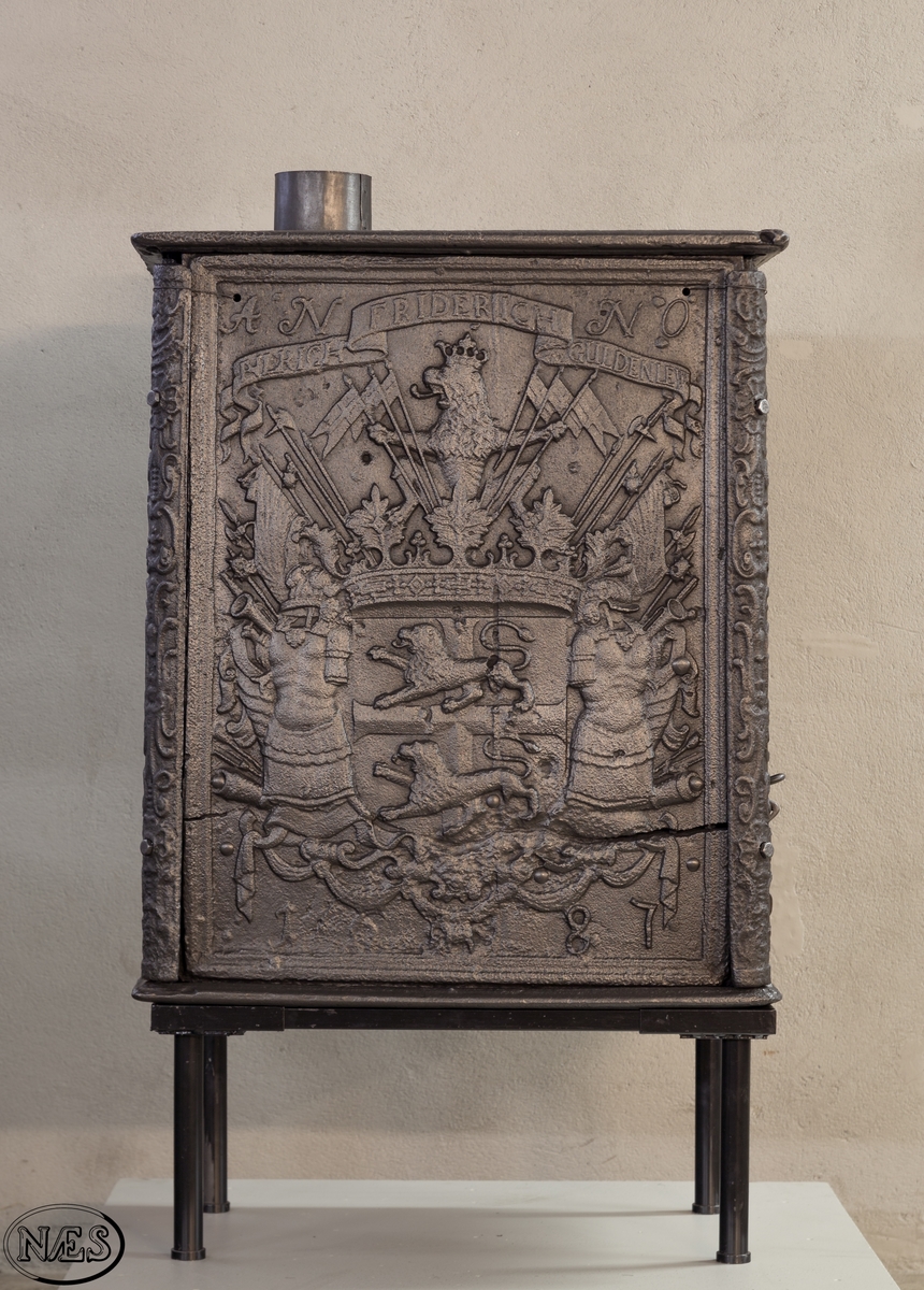 Kasseovn med ovnsdør på kortsiden og røykuttak i topplaten. Ovnen har løse skinner med bruskornamenter. På langsidene er Gyldenløves våpen omgitt av krigerske emblemer med innskrift i knekket bånd over. Øverst står det innskrevet ANNO og nederst på platen 1687. På kortsiden er det øverst krigerske emblemer og nederst den norske løve omkranset av brusk-kartusj.