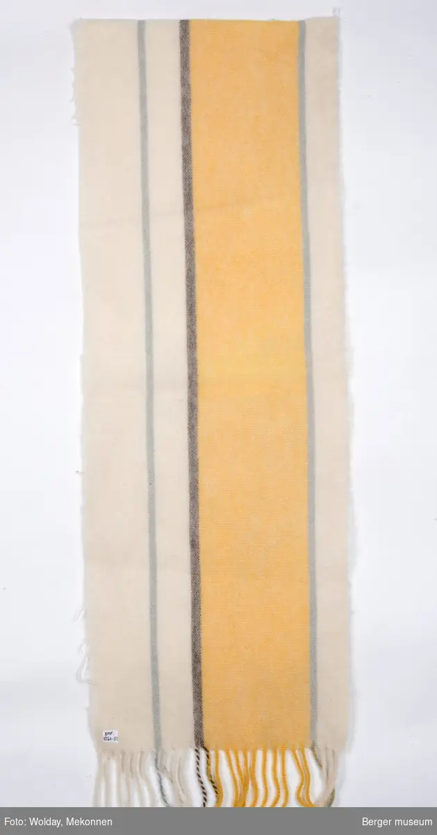 En avlang pleddprøve med stripemønster langs lengderetning; et bredt gult felt er adskilt av grå striper på hver side, og har hhv to og ett stripefelt i offwhite på hver side. Den ene lyse stripefeltet er delt av en smal grå stripe. Prøven har klippede kanter, og frynsekant på ene kortenden.