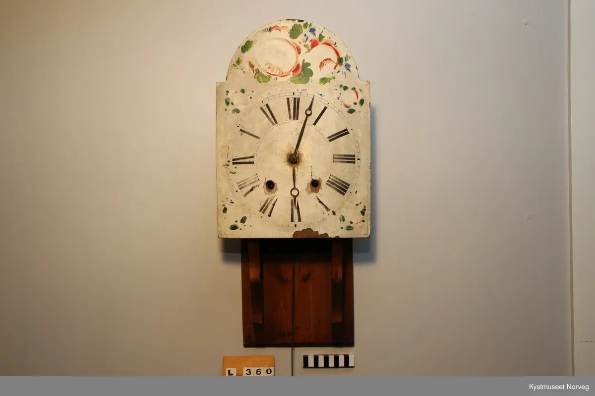 Veggur med 8-dagers verk. Hvitmalt urskive med romertall. Veggklokka er utført i heltre. Urskiva er dekorert med blomstermotiv i fargene rødt,grønt, bått og svart