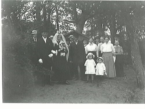 Brupar och bröllopsgäster i en träddunge med en bod i bakgrunden. Bruden klädd i svart klänning, vit slöja och brudkrona. Brudgummen klädd i frack och hög hatt. Två barn i blomprydda hattar står framför de vuxna gästerna.