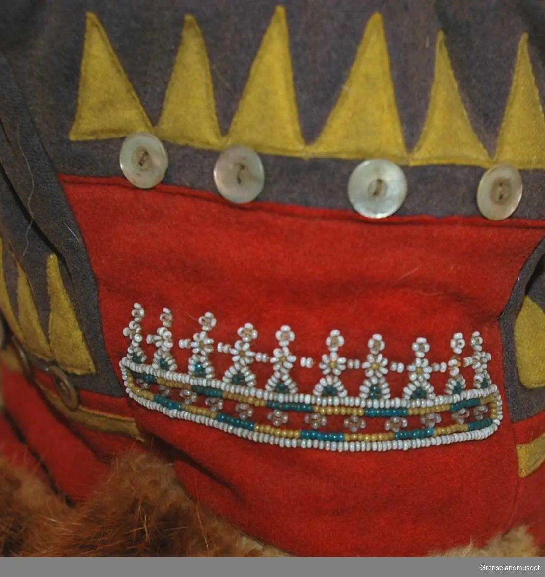 Av blå og rød klede med gul dekor (zik zak bånd) av klede. 12 hvite knapper og perlebroderi . Foret med reveskinn
