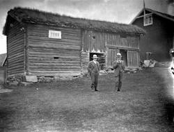 To menn ved tømret uthus på gardstun.