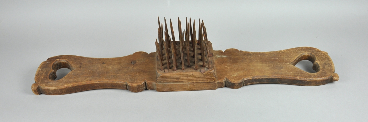 Linhekle av tre med hull som hjerter i hver ende av brettet. På midten er det festet en trekloss med stålspiker.