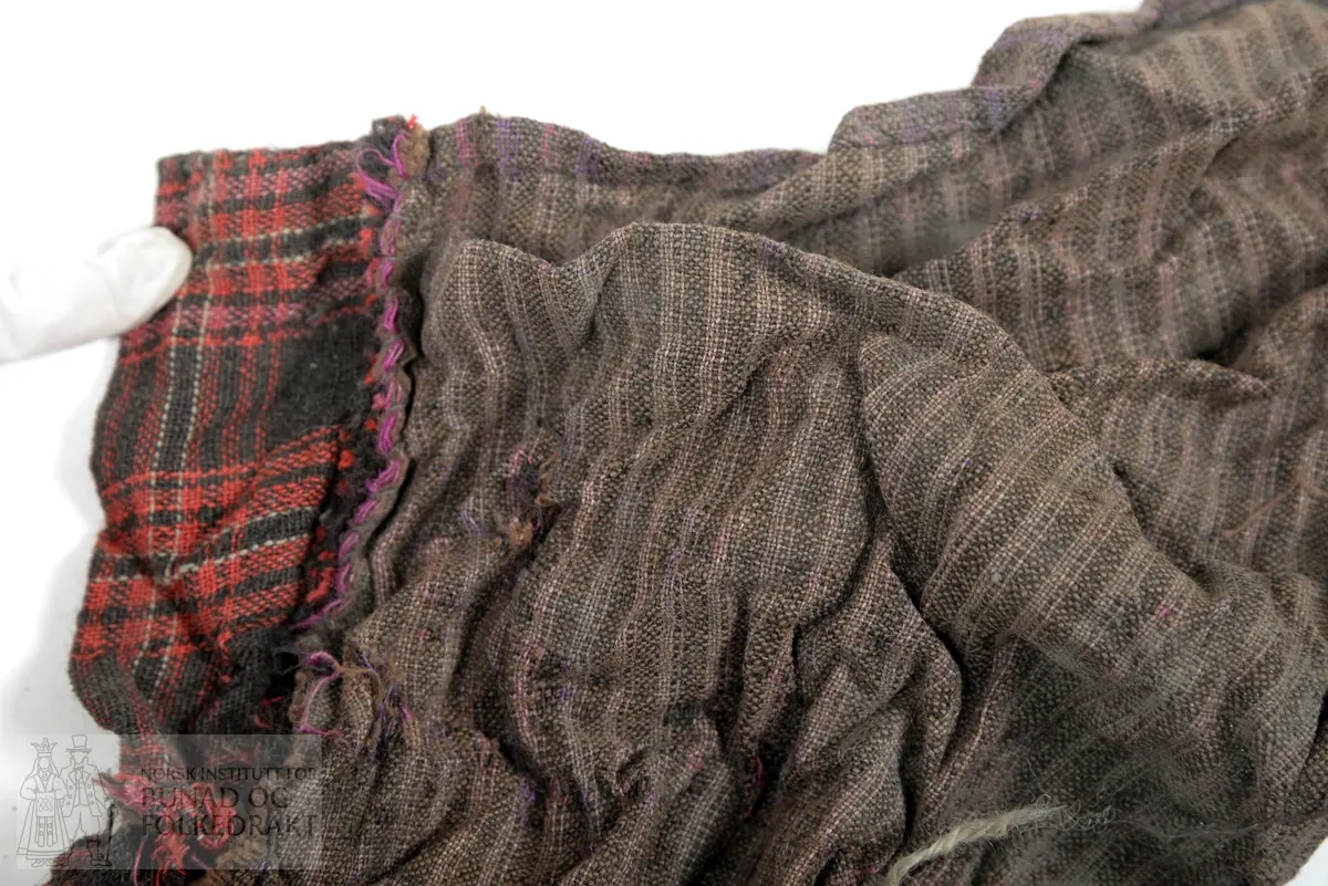 Svært utslitt del av ein stakk med linning, nederkant og ei metallhekte i eine enden av linning. Nedanfor denne delen av linning er det sauma fald som truleg er splitten midt framme på stakken. Av tre ulike verkensstoff, lerretsbinding med bomull i renning, ull i innslag. Stripete stoff i brun og lilla, svært utydeleg lillafargar. Fleire små hol etter mus, har vore nytta som stappefille.