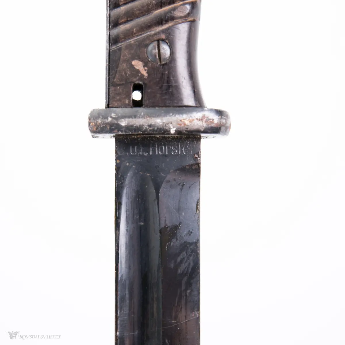 Metallhåndtak med lang pommel og geværfeste med fjæret knapp i bakkant. Serratert tregrep som er festet med 2 skruer. Langt spisst stålblad med egg og blodrand. Metallslire med kule enden og krok for festing til grunnutrustning/belte.