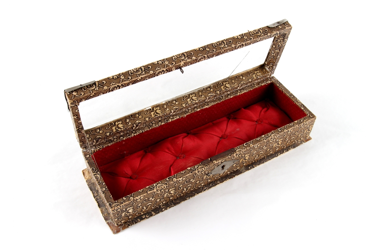 Avlangt smykkeskrin med hengsler og lokk. Lås og hjørnebeslag i metall. Lokket har et glassvindu. Foret med tekstil. Brukt til oppbevaring av små smykkegjenstander og en liten nøkkel.