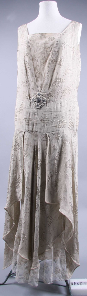 AV grålig voile brochert med sølv i et mønster av tynne grener med blader og klaser med bær på kryss og tvers over hele stoffet slik at baksiden blir nesten helt dekket av sølvtråder. Kjolen har sidt liv med innfelt, bredt hoftestykke som poser litt i underkanten. Hoftestykket har søm midt foran og er festet sammen bak, men fortsetter i to lange ender som faller helt ned til kjolekanten. Livet er utringet i en dyp spiss både foran og bak. Nederste del av spissen fylles ut av underkjolen av gråhvit crepe-du-chine, sekundært overtrukket med en fin silkeknipling med broderte blomster av sølvtråd. Kjole og underkjole er nå nestet sammen langs utringingen, underkjolen går litt lengre opp foran enn bak. Kjolen er ermeløs, for- og bakstykke møtes i en 5 cm. bred stropp over skulderen. skjørtet består av en forbredd og en bakbredd tatt på skrå av stoffet, utskrådd i sidene og sterkt buet i underkanten og litt lengre foran enn bak. For- og bakbredd møtes ikke i sidene før 15,5 cm. fra underkanten hvor de er sydd sammen med en smal strimmel av stoffet, øverst 4 cm, nederst 1 cm. bred. Mellomrommet i sidene dekkes av paneler, utskrådd som for- og bakbredden og tatt på skrå av stoffet. Underkanten er buet, nokså spiss på midten. Kanten nedentil, rundt panelene, hals- og ermutringingen avsluttes med en rullor av grålig voile. Blylodd i beltespissene og spiss foran. 
Kjolen er antagelig nokså snart blitt forlenget med et bredt stykke silkeknipling med broderte blomster av sølvtråd. Kniplingen er bredest på sidene, ca. 30 cm, men smalner til 2 cm. foran og 6 cm. midt bak. Samtidig er underkjolen i utringingen foran og bak blitt trukket med samme slags knipling, og underkjolen selv forlenget med et innfelt stykke fra livet og nedover, 41,5 cm. og en kile satt inn på hver side nederst.
Ved spissen av utringingen foran er påsydd en agraman med similistener, sorte, korte stråperler og riflete sølvperler som imiterer possement-arbeide.