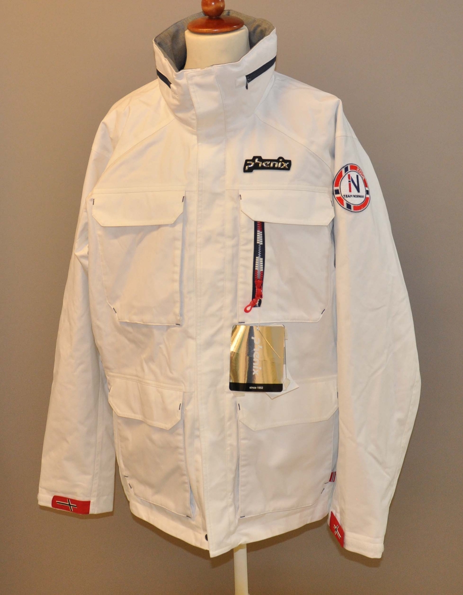 Hvit vinterjakke med glidelås og store lommer. Deltagermerke på venstre bryst og de olympiske ringer på ryggen. Mørkeblå glidelås i halsen for hette. Mørkeblått fleecefor med norsk flagg i ryggen.