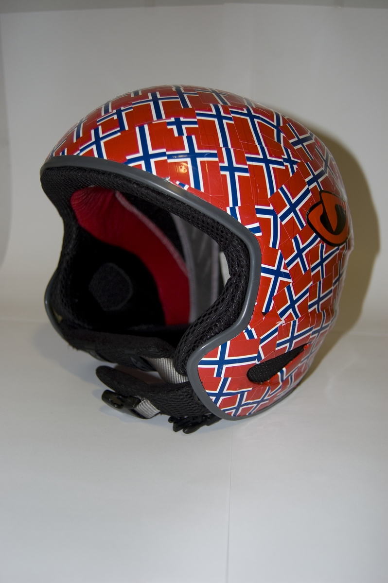 Alpinhjelm med påklistert merker med motiv av det norske flagget. Stramme- og tilpasningsanordning bak på hjelmen.