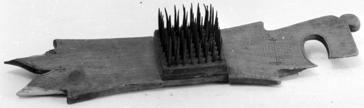 Hampehekle av tre med ovalt hull i den ene enden. Brettet har en trekloss på midten med stålnagler og er skadet i begge ender.