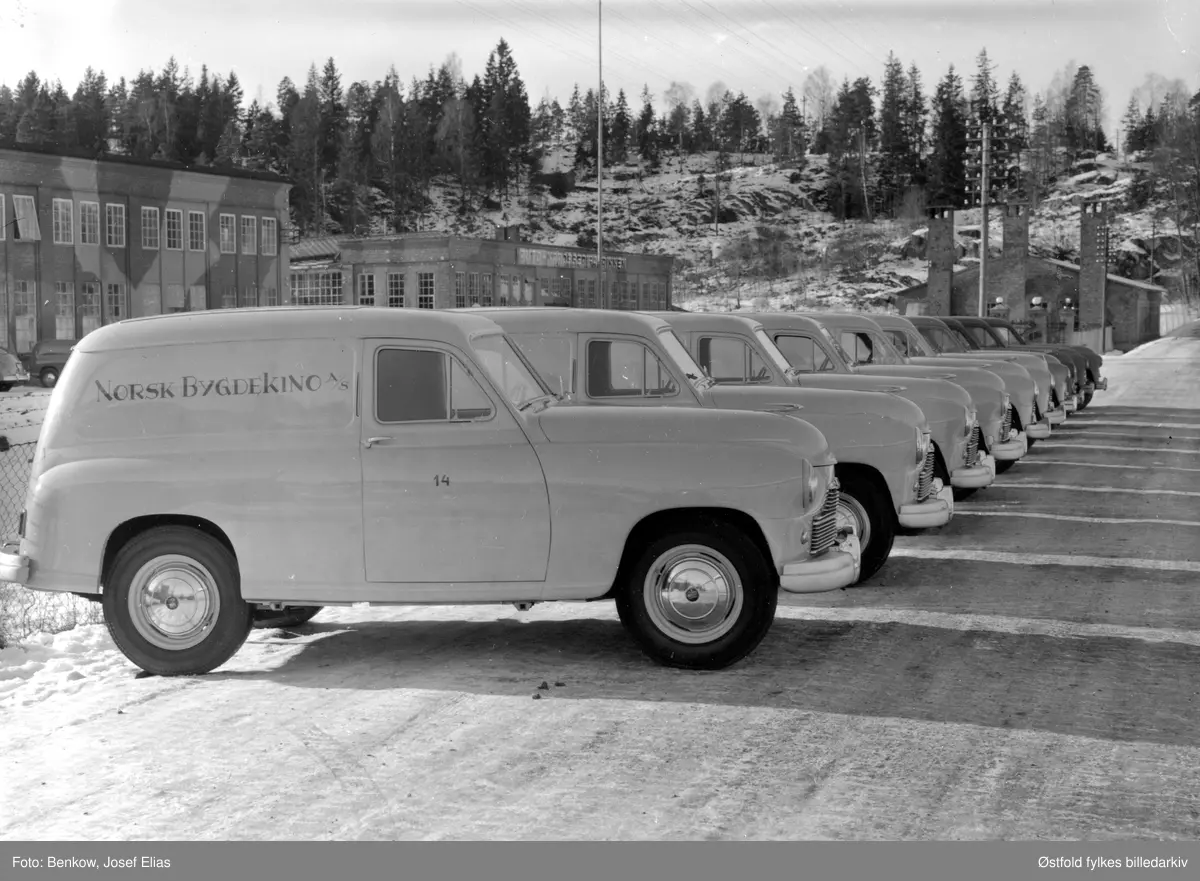 Nyproduserte biler til Norsk Bygdekino A/S utenfor Den Norske Automobilfabrikk på Kambo i Moss. Standard Vanguard ble klargjort, men ikke montert på Kambo. Oppstilt rekke av  gule varevogner til Norsk Bygdekino A/S som ble etablert 23. juli 1950. 
Fabrikkbygningen har fortsatt kamuflaskemaling fra 2. verdenskrig. 
Fabrikken ble rekvirert av tyskerne under 2. verdenskrig og ble brukt som verksted for tyske Junker-fly. 

Moss byleksikon: 
"Norsk Automobilfabrikk, Møllebakken 4, Kambo i Moss. Etablert i 1918. Her ble det produsert 77 lastebiler i løpet av tre år. Men så gikk bedriften konkurs i 1923. Den ble reetablert 1929 av Gustav Henning. Bedriften ble startet i samarbeid med svenske Scania-Vabis. De siste to årene ble det produsert karosserier og enkelte deler. Men 13 år senere - i 1936 - ble Den Norske Automobilfabrikk etablert, i de samme lokalene. Der ble det ikke produset biler, men karosserier til Plymouthbiler og Fargo lastebiler ble satt sammen. Bedriften kunne fremstille gjennomsnitlig ett karosseri om dagen. Det var stort sett lasteplan og førerhus som ble laget på fabrikken. Den totale produksjonen ble anslått til i underkant av 2200 biler.
Etter krigen ble fabrikken brukt til å klargjøre importerte biler. Virksomheten opphørte i 1963-64.