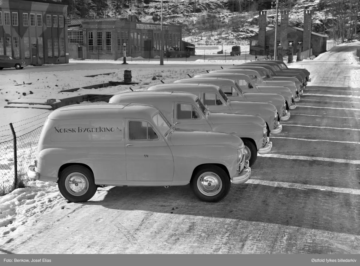 Nyproduserte biler til Norsk Bygdekino A/S utenfor Den Norske Automobilfabrikk på Kambo i Moss. Standard Vanguard ble klargjort, men ikke montert på Kambo. Oppstilt rekke av  gule varevogner til Norsk Bygdekino A/S som ble etablert 23. juli 1950. 
Fabrikkbygningen har fortsatt kamuflaskemaling fra 2. verdenskrig. 
Fabrikken ble rekvirert av tyskerne under 2. verdenskrig og ble brukt som verksted for tyske Junker-fly. 

Moss byleksikon: 
"Norsk Automobilfabrikk, Møllebakken 4, Kambo i Moss. Etablert i 1918. Her ble det produsert 77 lastebiler i løpet av tre år. Men så gikk bedriften konkurs i 1923. Den ble reetablert 1929 av Gustav Henning. Bedriften ble startet i samarbeid med svenske Scania-Vabis. De siste to årene ble det produsert karosserier og enkelte deler. Men 13 år senere - i 1936 - ble Den Norske Automobilfabrikk etablert, i de samme lokalene. Der ble det ikke produset biler, men karosserier til Plymouthbiler og Fargo lastebiler ble satt sammen. Bedriften kunne fremstille gjennomsnitlig ett karosseri om dagen. Det var stort sett lasteplan og førerhus som ble laget på fabrikken. Den totale produksjonen ble anslått til i underkant av 2200 biler.
Etter krigen ble fabrikken brukt til å klargjøre importerte biler. Virksomheten opphørte i 1963-64.