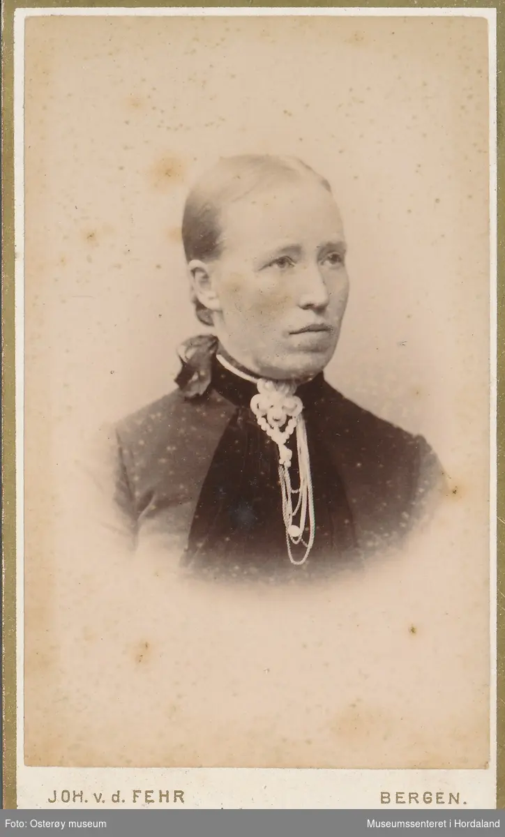 portrettfotografi av kvinne med midtskill og håret i knute i nakken. mørk bluse inne i ljosare jakke, sølje med kjeder i halsen