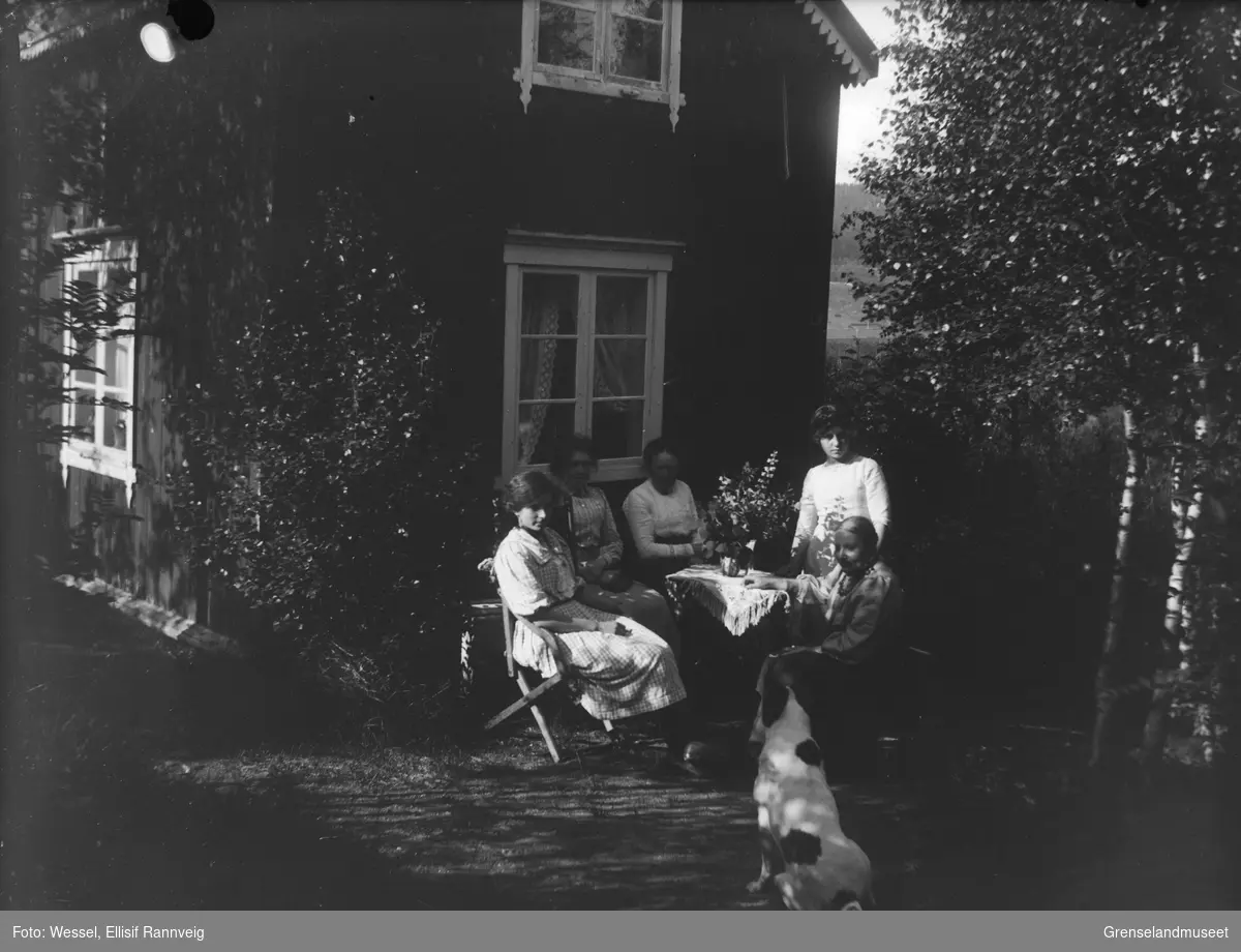 En gruppe kvinner fotografert sittende ved et bord ute i hagen, en hund i forgrunnen.