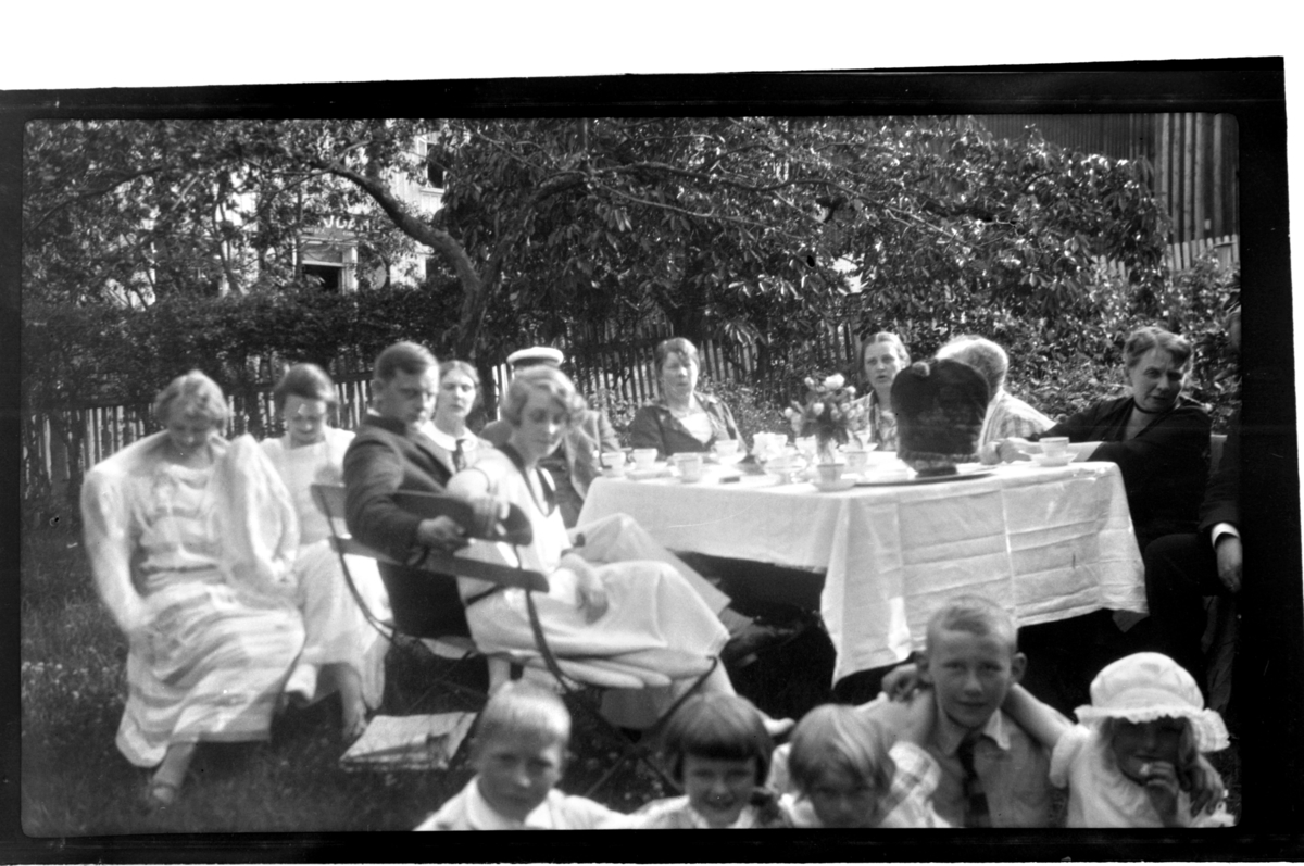 Voksne og barn samlet til teselskap rundt bord i hage i England. Edna og Percy Stark blant gjestene. Fotografert 1925.