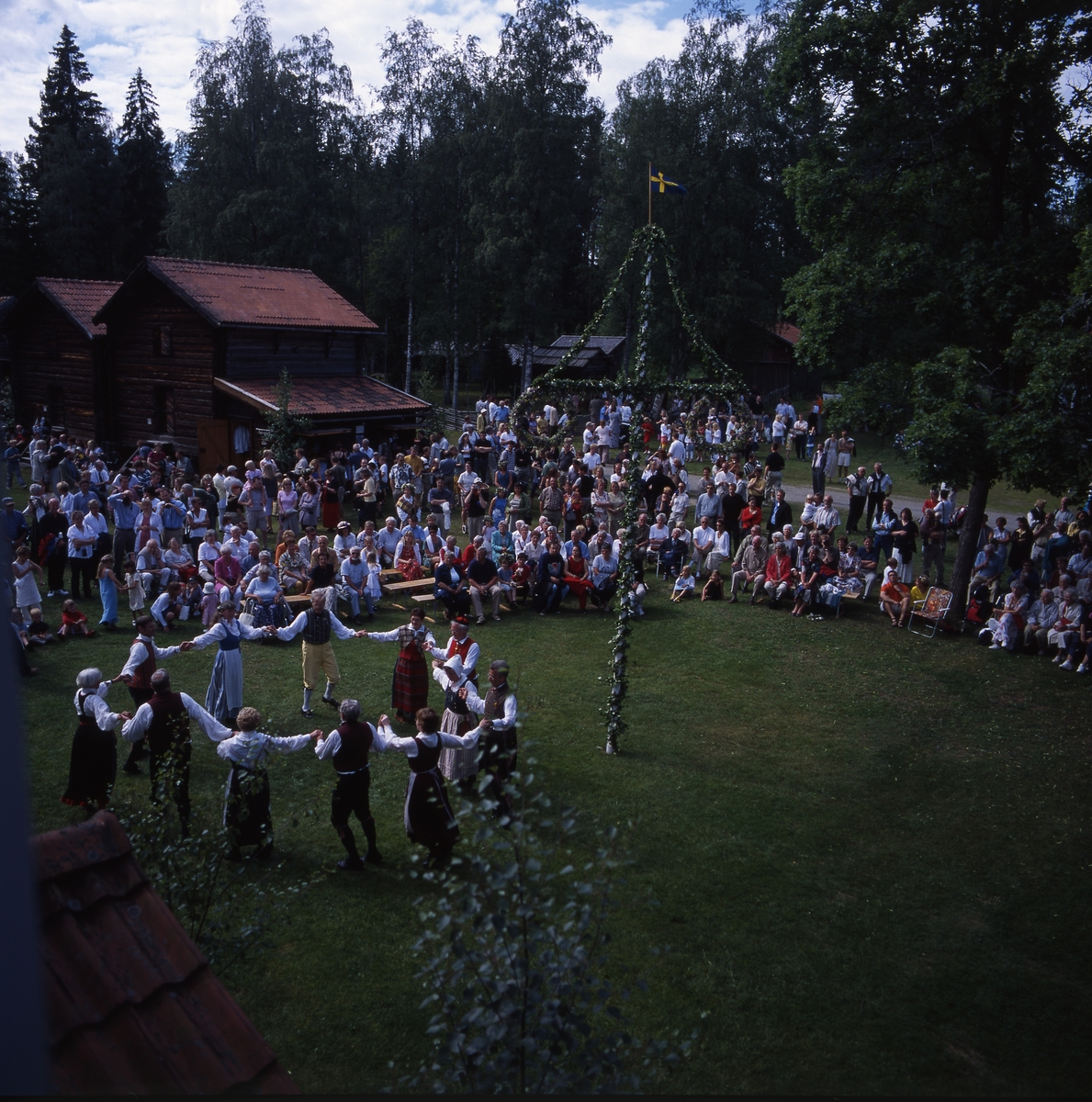 Midsommarfest vid Västerby hembygdsby i Rengsjö år 2000. Dansare i folkdräkt uppträder runt midsommarstången. En stor publik sitter på bänkar och ser på.