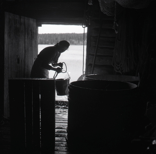 I dörren ser vi en person med hink i silhuett. Från fiskeläget i Norrfjärden Gnarp  3 juni 1954.