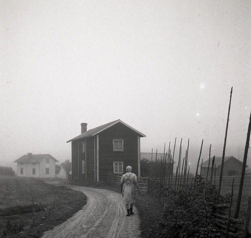 En kvinna vandrar på en grusväg mot några gårdar i dimma, Knåda, Edsbyn september 1949.