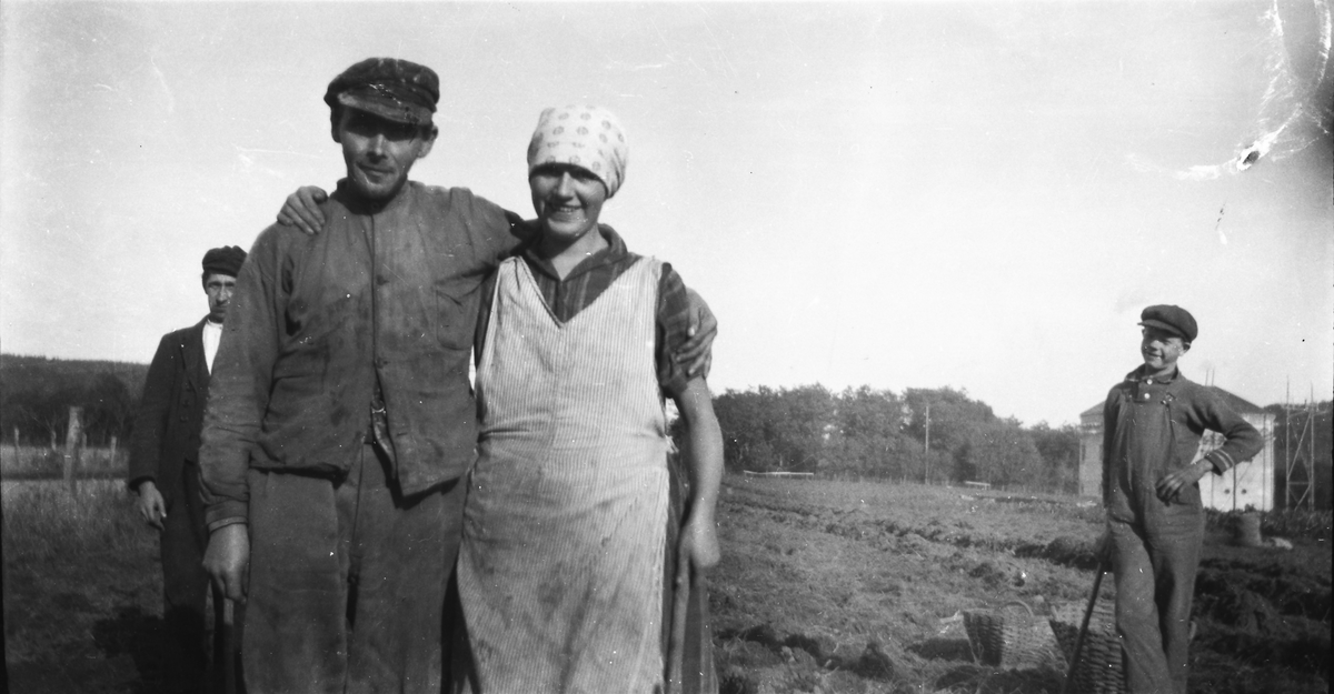 Kloar-Erik och Ellen i Kalvhult poserar på en potatisåker. I bakgrunden står en pojke och hänger på ett redskap bredvid ett par potatiskorgar. Bakom honom syns en kraftstation.