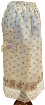Midjeförkläde av mönstrat bomullstyg i vitt med blommönster. Förklädet är rynkat i midjan, utan linning samt har rosa sidenband till knytband. Nertill avslutas förklädet med en maskintillverkad vit spets. Förklädet har en ficka med en rosa sidenrosett fastydd på utsidan.