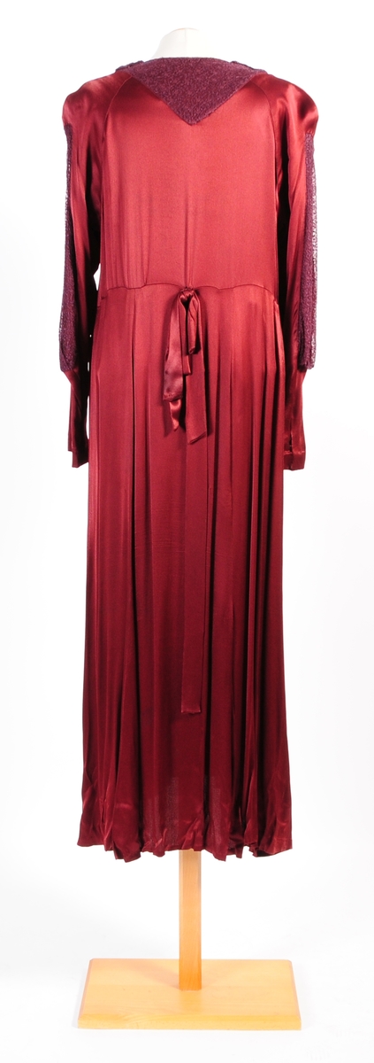 Lång klänning av vinfärgad crepe de chine. Klänningen har besättning av tyllspets i samma färg som på ärmarna och runt halsen.