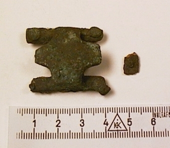 Spänne eller beslag, möjligen till bälte, av brons. Smycket har rester av nitplattor av järn på undersidan, fästa med fyra bronsnitar, en i vardera hörnet.
