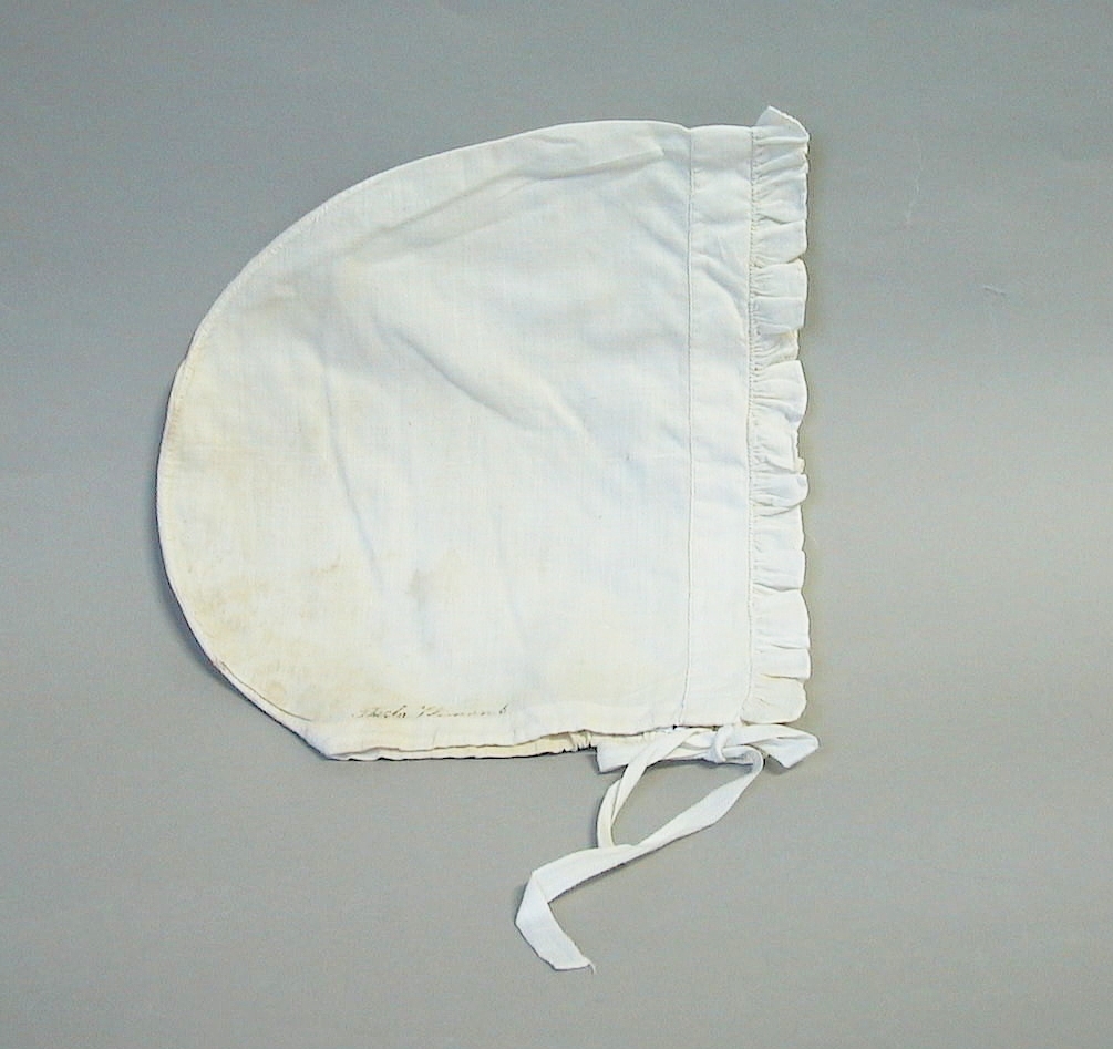 Nattmössa av vitt bomullstyg, sydd med en rynkad kappa efter framkanten. Längs nerkanten går en sydd kanal med ett iträtt vitt bomullsband.