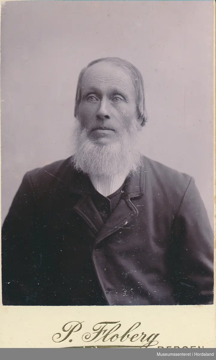halvfigurfotografi av mann med tynt hår og skjegg, kledd i mørk jakke med vest og kvit skjorte