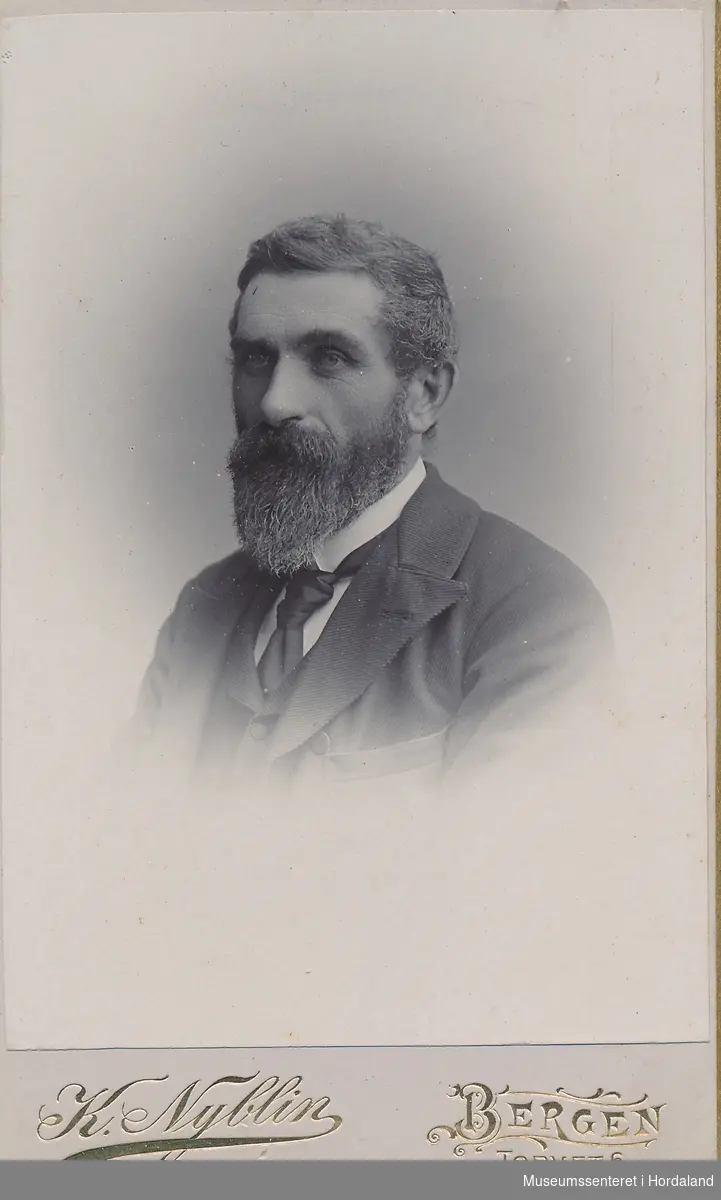 Portrettfotografi av mann med skjegg, mørk jakke og vest og kvit skjorte med mørkt slips.