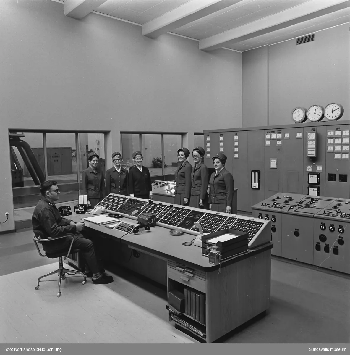 Sex kvinnor i uniform samt en man vid en manöverpanel på Bergeforsens kraftverk. Fotograferat för en affisch.