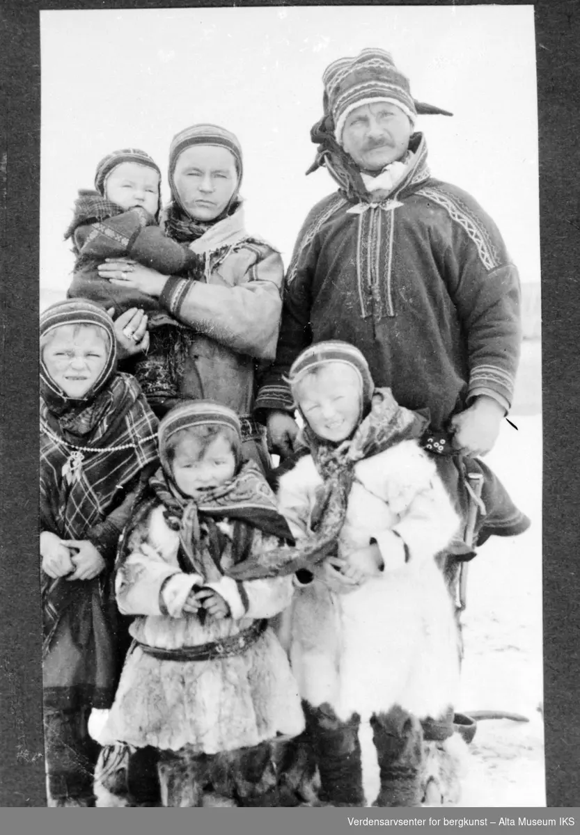 Bilde av en samisk familie på seks, mor holder den yngste og barna foran er kledd i reinskinn.