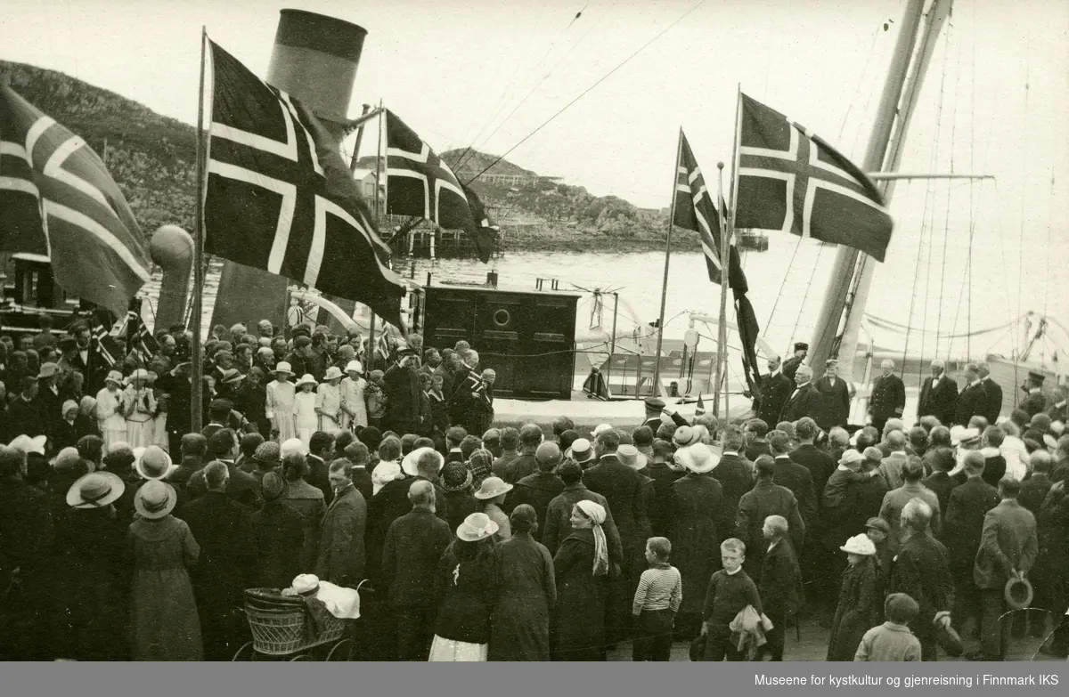 Postkort. Kongebesøk i Honningsvåg. Kong Haakon VII går i land fra "Heimdal" på dampskipskaia. Feststemte folk med flagg møter kongen. 28.06.1922.