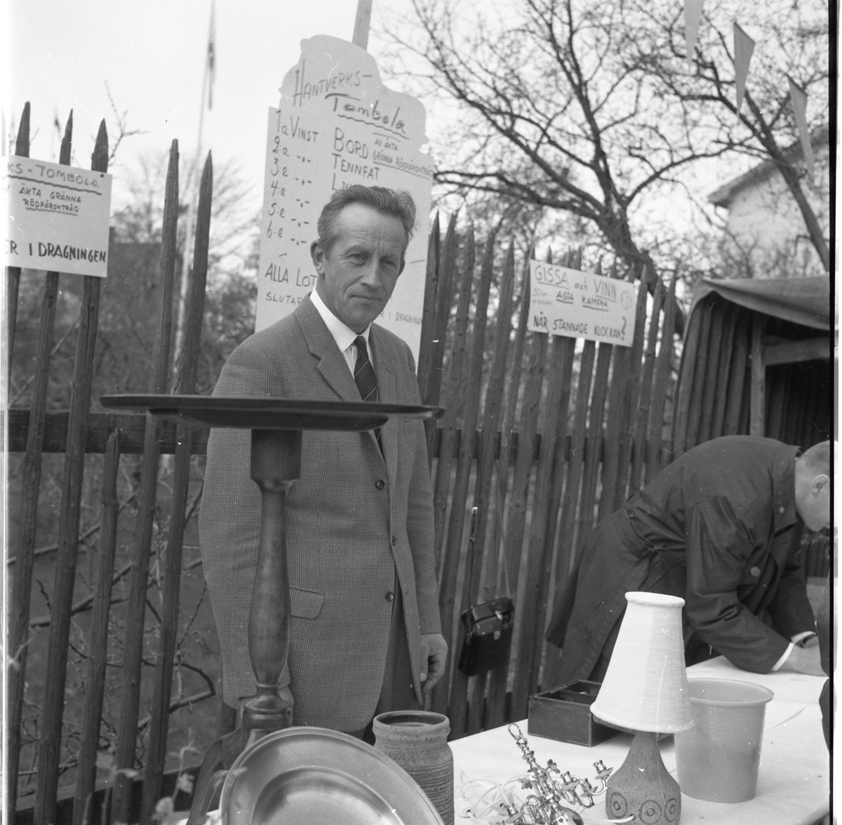 Lions vårmarknad 1964. Herbert Gustafsson står bakom ett tombola-bord på Sjögatan. Lotterivinsterna är hantverksprodukter. Första pris ett bord av äkta Gränna rödpäronträd.