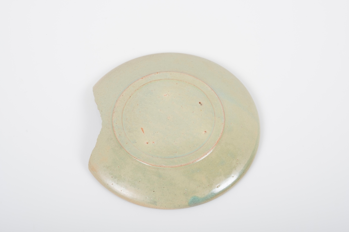 Skål i keramikk med grønn lasur. Spor etter tre knotter på bunnen, usikker funksjon. Bunnen har matt overflate. En brukket del av skålen mangler.