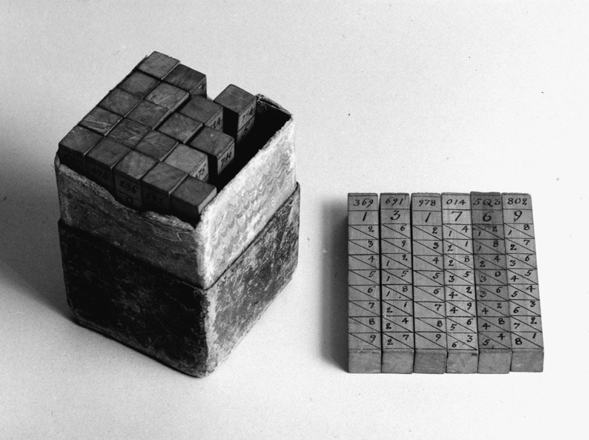 Napiers räknestavar för multiplikation av intill åttasiffriga tal med alla siffror av samma slag. Stavarnas diameter 13 x 13 mm. Komplett med 25 stavar i fodral av papp. Denna serie liknar mest den i katalogen "Mathematics I. Calculating machines and instruments", London 1926 under n:o 7, sid 11 upptagna, men samtliga multiplicandstavar är upptill på alla sidor försedda med siffror, som hänvisa till vilka siffror-multiplar, som befinner sig på övriga sidor. Dessutom finns kvadranterna och kuberna på talen 1-9. 25 st. neperska räknestavar (av trä med siffror i tusch) enligt John Napier (f. 1550, d. 1617, skrev vanligen Neper), för multiplikation av 8-11-siffriga tal med alla siffror av samma slag eller högst 24-siffriga tal. En stav med sifforna 1-9 i var sin vågrät rad på endast en sida bör vid räkning läggas till vänster om stavarna för att undvika förväxling av multipelrad vid produktens avläsning. På motsatta sidan av femte staven fr. v. på fotot finns kvadrater för talen 1-9. Likaså finns en stav med kuber för talen 1-9 på en sida. Stavarnas längd: 90 mm, tvärsnitt 13 x 13 mm. Den i katalog fr. Science Museum uppgivna räknevalsen saknar hänvisning upptill på varje stav till de på dennas övriga sidor befintliga siffrorna med multiplar. Antalet stavsidor med multiplar för vardera talen 0-9: 0  1  2  3  4  5  6  7  8  9 så att multiplikanden kan bestå av högst 10 8  9 10  8 10  10 9 11 11 = 94 sidor 8-11 siffror av samma slag.