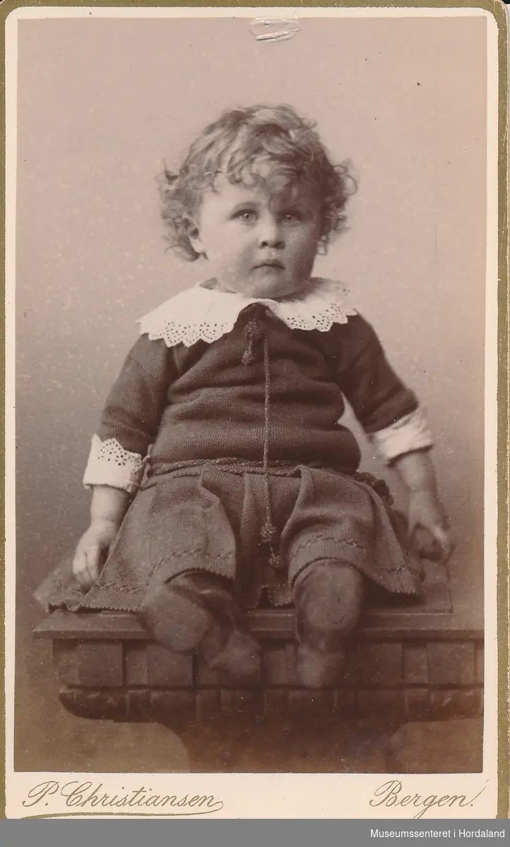 fotografi av ein liten gut med ljose krøllar og blondekrage/mansjettar som sit på eit bord