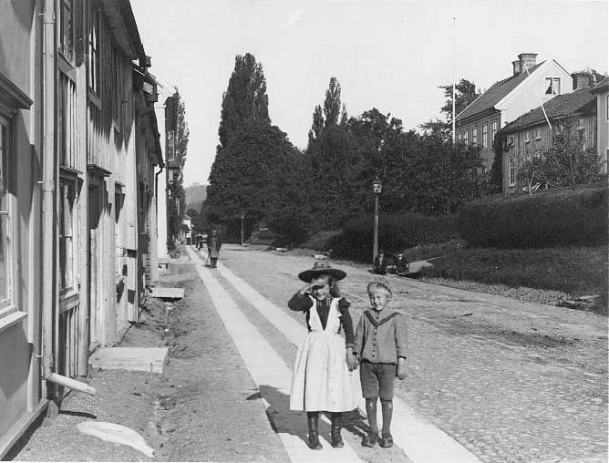 Brahegatan mot norr. Till höger syns Brahegatan 66 och 68. På trottoaren vända mot fotografen, och mot solen, står en flicka som håller en pojke i handen. Bakom dem går en med förskinn och på andra sidan gatan sitter två pojkar vid vägrenen.