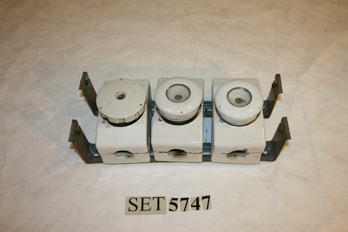 4 rektangulære boksar av kvitt porselen med ei sikring i kvar, fest til to jernbrakettar.