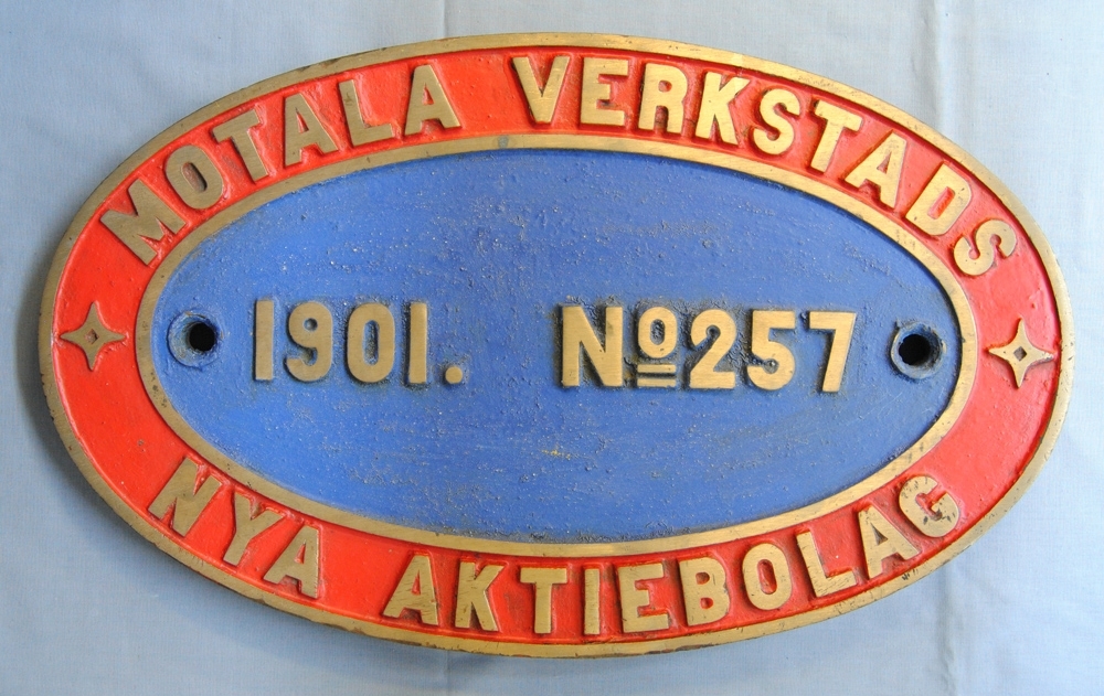 Oval loktillverkarskylt av mässing, målad i rött och blått med gula bokstäver. Text: "Motala verkstads Nya Aktiebolag. 1901. No. 257".

Modell/Fabrikat/typ: No 257, loknr. 18