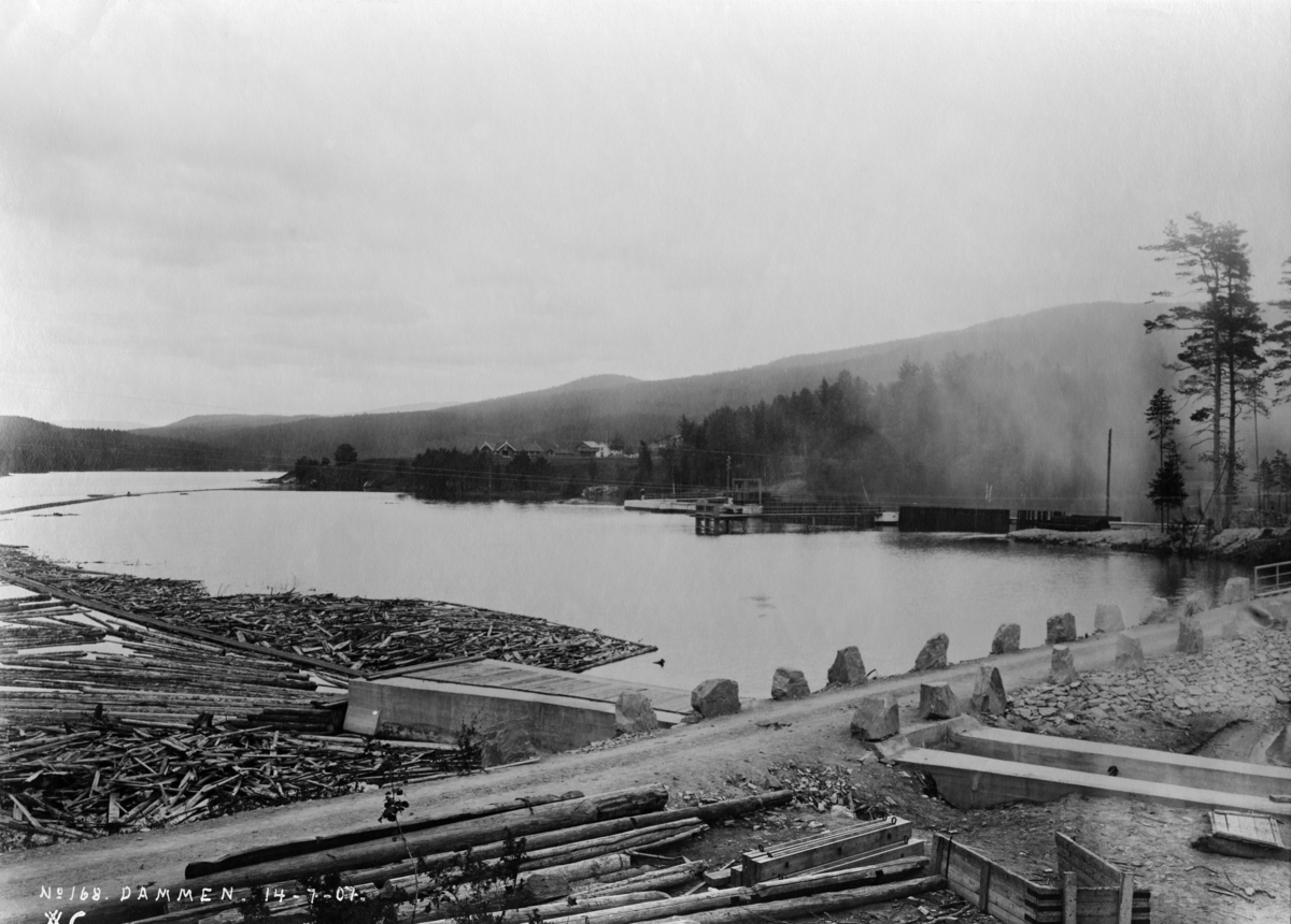 Inntaket til tømmerrenna ved fra Kloumannsjøen forbi Svelgfossen i Tinnåa i Telemark, fotografert i 1907.  Denne nye fløtingsleden ble innviet 14. mai dette året.  Den øvre delen av anlegget ble bygd av Norsk hydroelektrisk kvælstofaktieselskab - Norsk Hydro - med et tilskudd en gang for alle på 60 000 kroner fra Skiensvassdragets Fellesfløtingsforening.  Her var innløpet støpt i betong.  Det førte mot en 450 meter lang tunnel i fjell, som var foret med tre, for å redusere faren for at tømmerstokkene skulle kile seg fast mot ujevnheter i berget.  Nedenfor tunnelen fortsatte Svelgfossrenna som en trekonstruksjon.  Denne parsellen var det Fellesfløtningsforeningen som finansierte.  Hele renna var 1 230 meter lang.  På dette fotografiet ser vi inntakspartiet - betongrenna - og vegen over damkrona med sine stabbesteiner i forgrunnen.  Til venstre ser vi dessuten litt av den V-formete lensa som ledet tømmeret fra den oppdemte Kloumannsjøen i Tinnåa mot tømmerrenna.  Utbyggingsarbeidet og oppdemminga som fulgte med hadde medført at betydelige arealer ble avskoget.  Hogstavfallet fløt også mot renna, noe som skapte en del startvansker.  Når dette problemet var ryddet av vegen, ble det påstukket 15 - 1600 tylfter tømmer - fordelt på to arbeidsskift - hvert døgn i høysesong. 