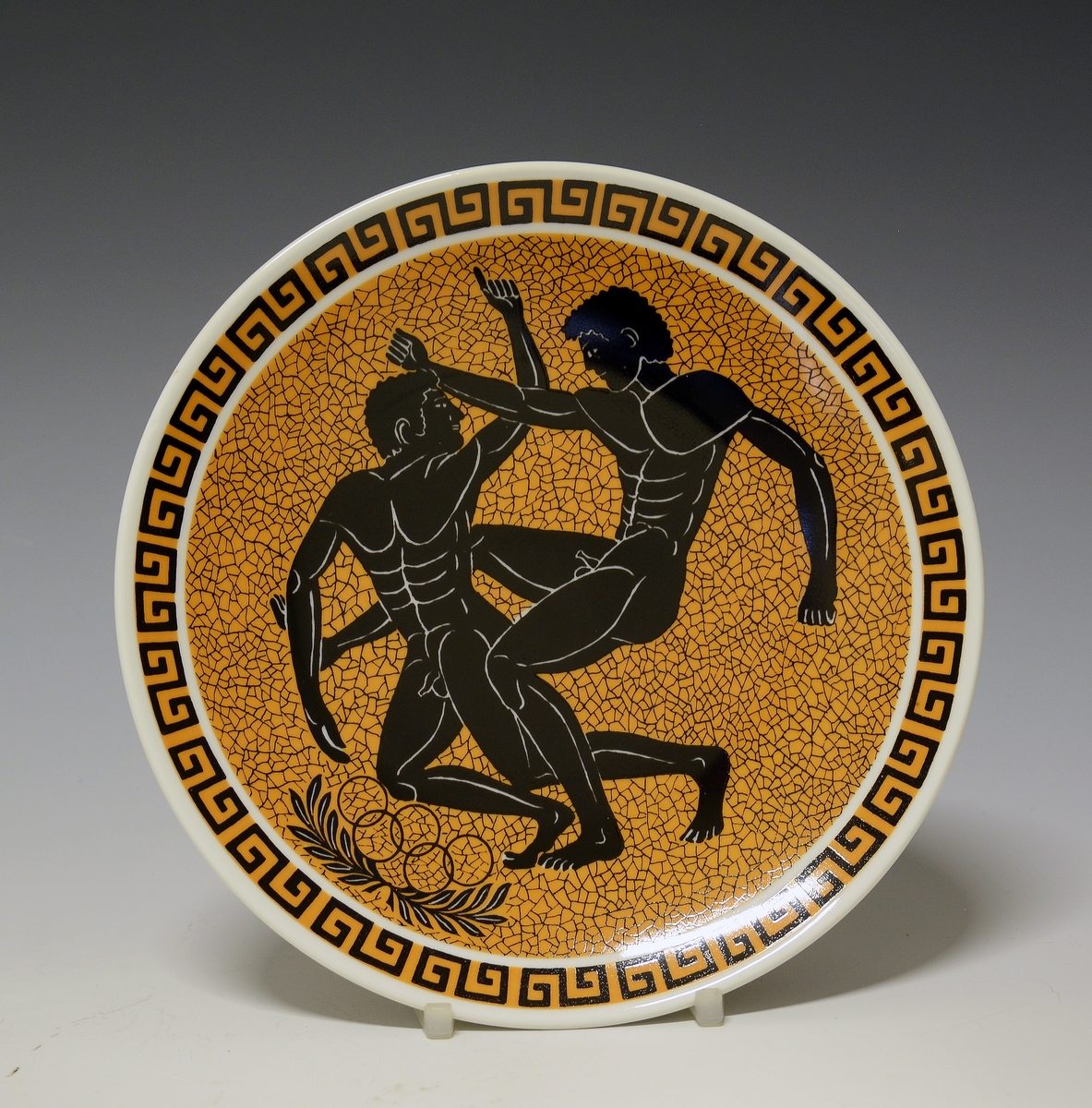 Platte med antikt gresk motiv, to nakne menn i en tvekamp kalt Pancratium. Under mennene de olympiske ringer på laurbær. Rundt fanen meanderbord.