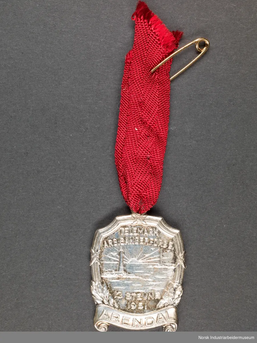 Medalje festet til rødt bånd. Festes med sikkerhetsnål. Motiv av fyrtårn i soldnedgang.