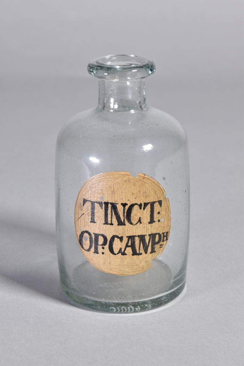 Flaska av klart glas, cylindrisk, kort hals med utsvängd mynning. Påmålad etikett med svart text.