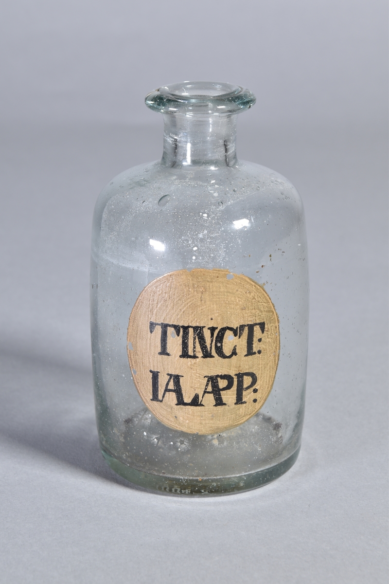 Flaska av klart glas, cylindrisk, kort hals med utsvängd mynning. Påmålad etikett med svart text. Rester av innehåll.
