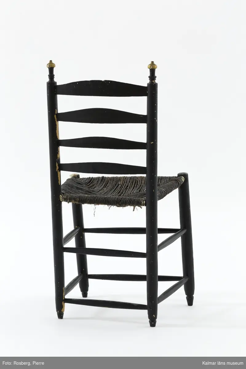 KLM 18989:4. Stol, stegstol, stegryggstol. Av typen Ladderback chair - stegryggstol. Av trä. Ryggstöd med fyra bladformade och tvärgående ryggbrickor. Ryggstolparna avslutas med snidade detaljer. Raka ben med dubbla rader av runtgående benslåar. Målad i svart med detaljer i guld. På ett flertal ställen syns att stolen varit målad i ljust gul, troligen originalfärg. Flätad sits av rotting i tre lager.