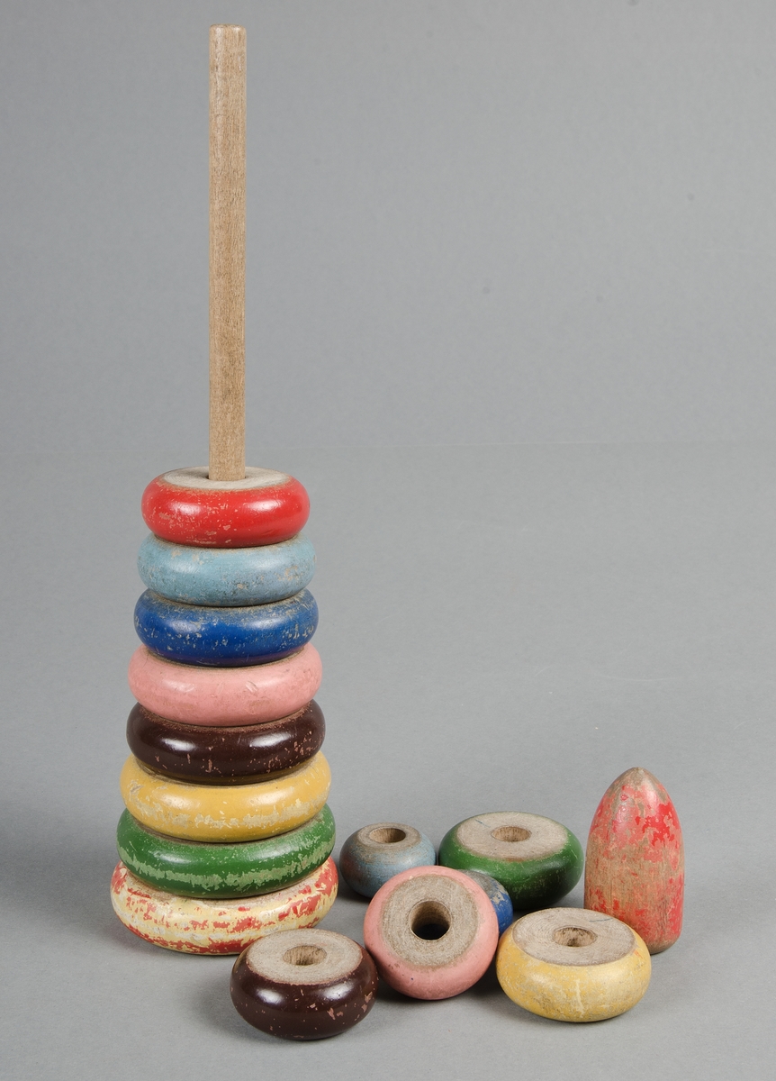 Träleksak i form av en pinne med träringar i olika storlek målade i olika färger, rött, grönt brunt, rosa, blått. Röd knopp. Underplatta som pinnen skall sitta i saknas.
