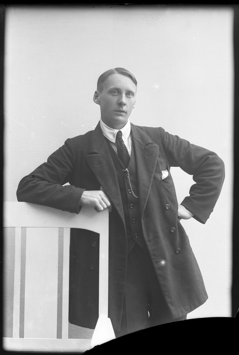 Porträtt av Karlström, iklädd tredelad kostym, vit skjorta och slips, lutad mot en stol.