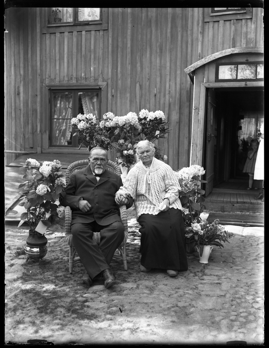 Janne Thorsell sitter ute i trädgården i en korgstol, omgiven av blommor på sin 70årsdag. Bredvid honom sitter hans fru (förmodligen Emma Christina Orrbin) och i dörröppningen bakom dem står en liten flicka.